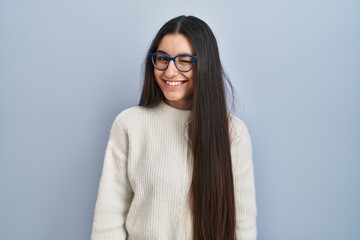 Young hispanic woman wearing casual sweater over blue background winking looking at the camera with...