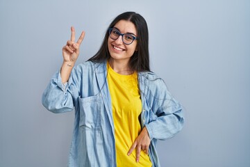 Young hispanic woman standing over blue background smiling looking to the camera showing fingers doing victory sign. number two.