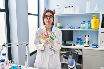 Young hispanic woman working at scientist laboratory holding money banknotes puffing cheeks with...
