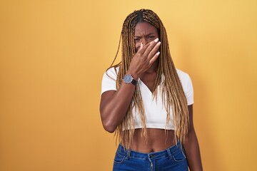 African american woman with braided hair standing over yellow background smelling something stinky and disgusting, intolerable smell, holding breath with fingers on nose. bad smell