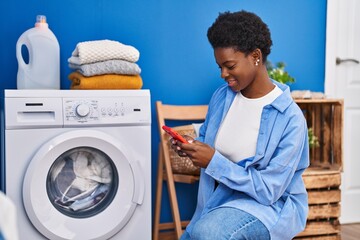 African american woman using smartphone waiting for washing machine at laundry room