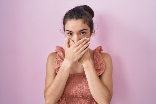 Young teenager girl standing over pink background shocked covering mouth with hands for mistake. secret concept.