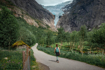 Woman hiking along path to Briksdalsbreen glacier in the mountains of Jostedalsbreen national park in Norway, green meadows with flowers in foreground - 559151688