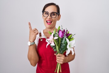 Middle age brunette woman wearing apron working at florist shop holding bouquet pointing finger up with successful idea. exited and happy. number one.