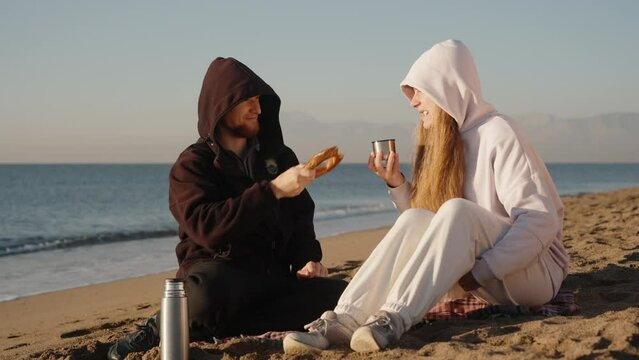 The couple arranged tomorrow on the seashore, It's cool and they are dressed in a black and white hoodie, they take turns eating a bagel and drinking tea.
