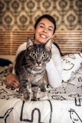 Portrait of a beautiful gray cat looking into the camera. Young woman plays with the cat on the bed. - 559152629