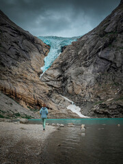 Naklejka premium Woman hiking along beach of glacier lake at Briksdalsbreen glacier in the mountains of Jostedalsbreen national park in Norway, turquoise water and blue ice