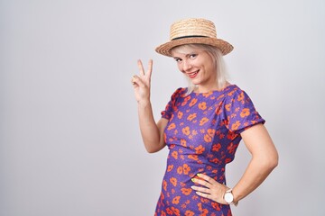 Young caucasian woman wearing flowers dress and summer hat smiling looking to the camera showing fingers doing victory sign. number two.