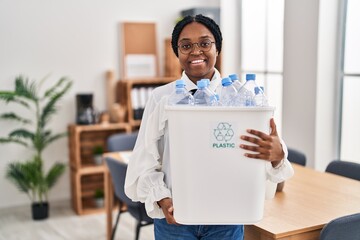 African american woman working at the office holding plastic bottle for recycling smiling with a...