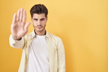 Young hispanic man standing over yellow background doing stop sing with palm of the hand. warning...