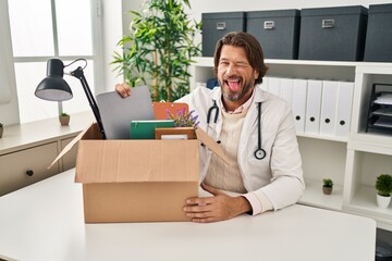 Handsome middle age doctor man holding box with items sticking tongue out happy with funny expression. - obrazy, fototapety, plakaty