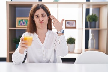Brunette woman drinking glass of orange juice shooting and killing oneself pointing hand and fingers to head like gun, suicide gesture.