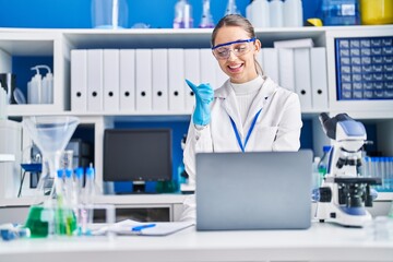 Young blonde woman working at scientist laboratory pointing thumb up to the side smiling happy with open mouth