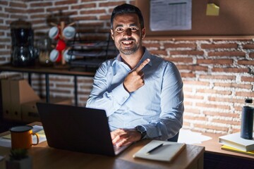 Hispanic man with beard working at the office at night cheerful with a smile on face pointing with hand and finger up to the side with happy and natural expression