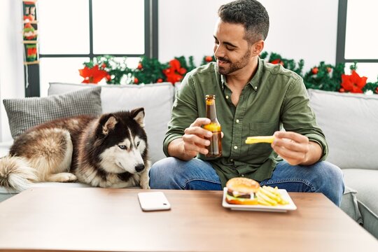 Young Hispanic Man Eating Hamburger Sitting On Sofa With Dog By Christmas Decor At Home