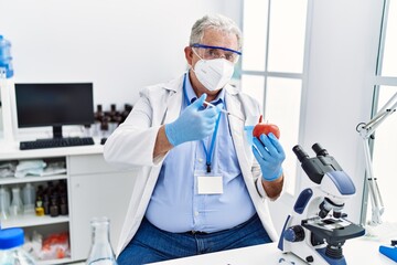 Middle age grey-haired man wearing scientist uniform holding syringe and apple at laboratory