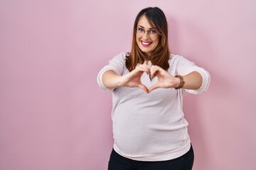 Pregnant woman standing over pink background smiling in love doing heart symbol shape with hands. romantic concept.