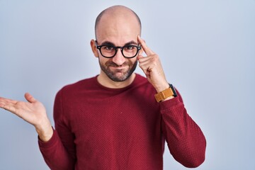 Young bald man with beard standing over white background wearing glasses confused and annoyed with open palm showing copy space and pointing finger to forehead. think about it.