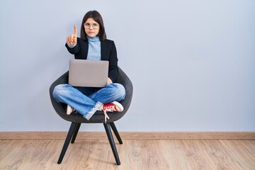 Young hispanic woman sitting on chair using computer laptop pointing with finger up and angry expression, showing no gesture