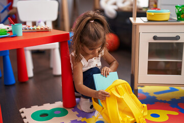 Adorable hispanic girl preschool student sitting on floor holding book of backpack at kindergarten