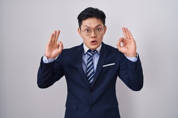 Young asian man wearing business suit and tie looking surprised and shocked doing ok approval symbol with fingers. crazy expression