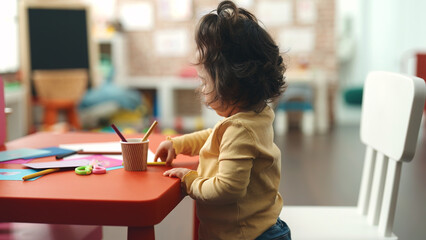 Adorable hispanic girl student sitting on table holding pencil color at kindergarten