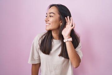 Young hispanic woman standing over pink background smiling with hand over ear listening an hearing to rumor or gossip. deafness concept.