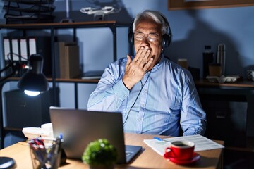 Hispanic senior man wearing call center agent headset at night bored yawning tired covering mouth...
