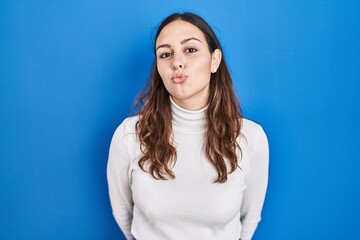 Obraz premium Young hispanic woman standing over blue background looking at the camera blowing a kiss on air being lovely and sexy. love expression.