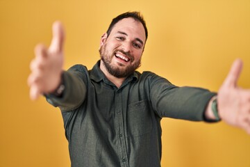Plus size hispanic man with beard standing over yellow background looking at the camera smiling with open arms for hug. cheerful expression embracing happiness.