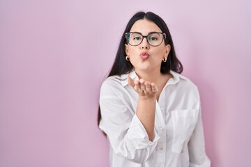 Young brunette woman standing over pink background looking at the camera blowing a kiss with hand on air being lovely and sexy. love expression.