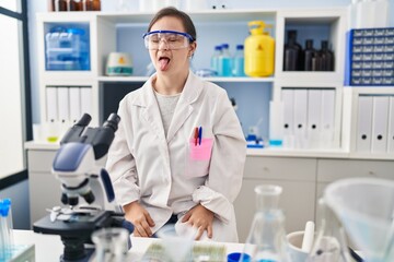Hispanic girl with down syndrome working at scientist laboratory sticking tongue out happy with funny expression. emotion concept.