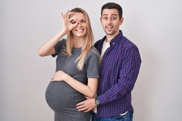 Young couple expecting a baby standing over white background doing ok gesture with hand smiling,...