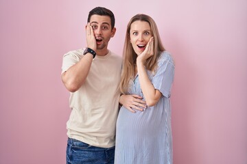 Naklejka premium Young couple expecting a baby standing over pink background afraid and shocked, surprise and amazed expression with hands on face