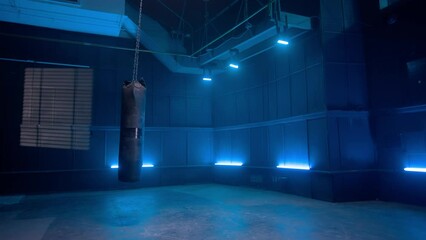 Long black punchbag hanging and swinging from chain in dark empty gym with blue neon lights. Leather punching bag for kickboxing, boxing, kicking and punching. Slow motion ready, 4K at 59.94fps.