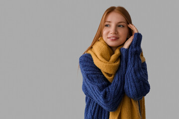 Young redhead woman in warm sweater and scarf on grey background