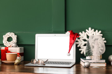 Laptop with Santa hat and Christmas decor on table near green wall