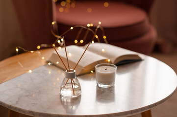 Cozy home atmosphere with liquid fragrance in glass bottle and sticks with open paper book over glow lights on marble table in living room close up. Winter holiday season.