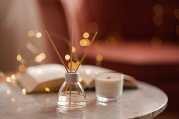 Cozy home atmosphere with liquid fragrance in glass bottle and sticks with open paper book over glow lights on marble table in living room close up. Winter holiday season.