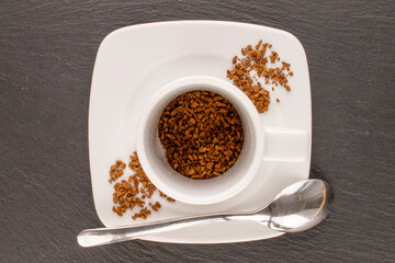 Fragrant instant coffee on white saucer with white cup, metal spoon on slate stone, macro, top view.