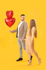 Happy young couple with heart-shaped balloons on yellow background. Valentine's Day celebration
