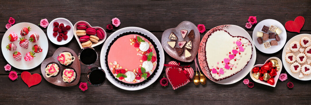 Valentines Day table scene with assorted desserts and sweets. Overhead view on a dark wood banner background. Love and hearts theme.
