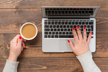 Top view of office desk table. Woman with red nails holding cup of coffee and working on modern laptop on wooden table