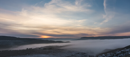 Winter panorama at sunset with snow covered hills with low fog in the valley and cloud cover, hills with snow and trees in winter at sunset © Denis