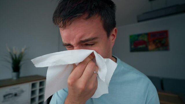 Irritated man sneezes trying to wipe running nose with napkin. Young male person caught cold on autumn day sitting in unheated apartment closeup