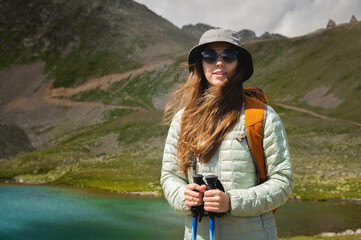 Naklejka premium Portrait of a smiling woman resting during a mountain hike. A woman with a backpack enjoys the view, standing at the lake and at the foot of the mountain