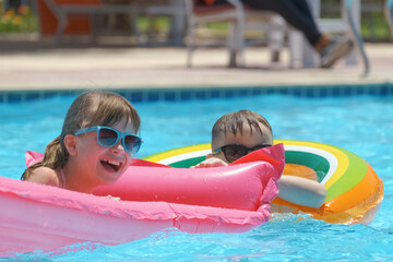 Naklejka premium Young joyful children boy and girl having fun swimming on inflatable air mattress in swimming pool with blue water on warm summer day on tropical vacations. Summertime activities concept