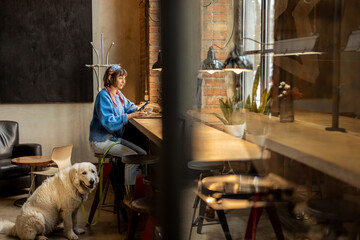 Woman sits with her cute adorable dog at modern coffee shop and works on laptop. Pet friendly places and modern digital lifestyle concept