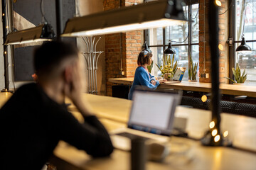 Woman works on laptop while sitting by the window at modern coffee shop. Wide view from the backside with people in front