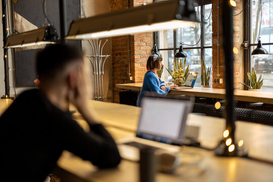 Woman Works On Laptop While Sitting By The Window At Modern Coffee Shop. Wide View From The Backside With People In Front